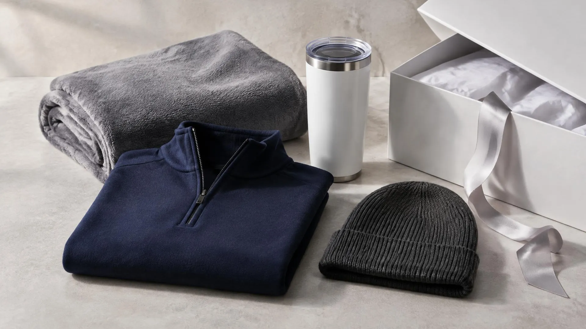 Folded navy quarter zip, gray fleece blanket, white insulated tumbler and charcoal knit beanie beside an open white gift box with ribbon.