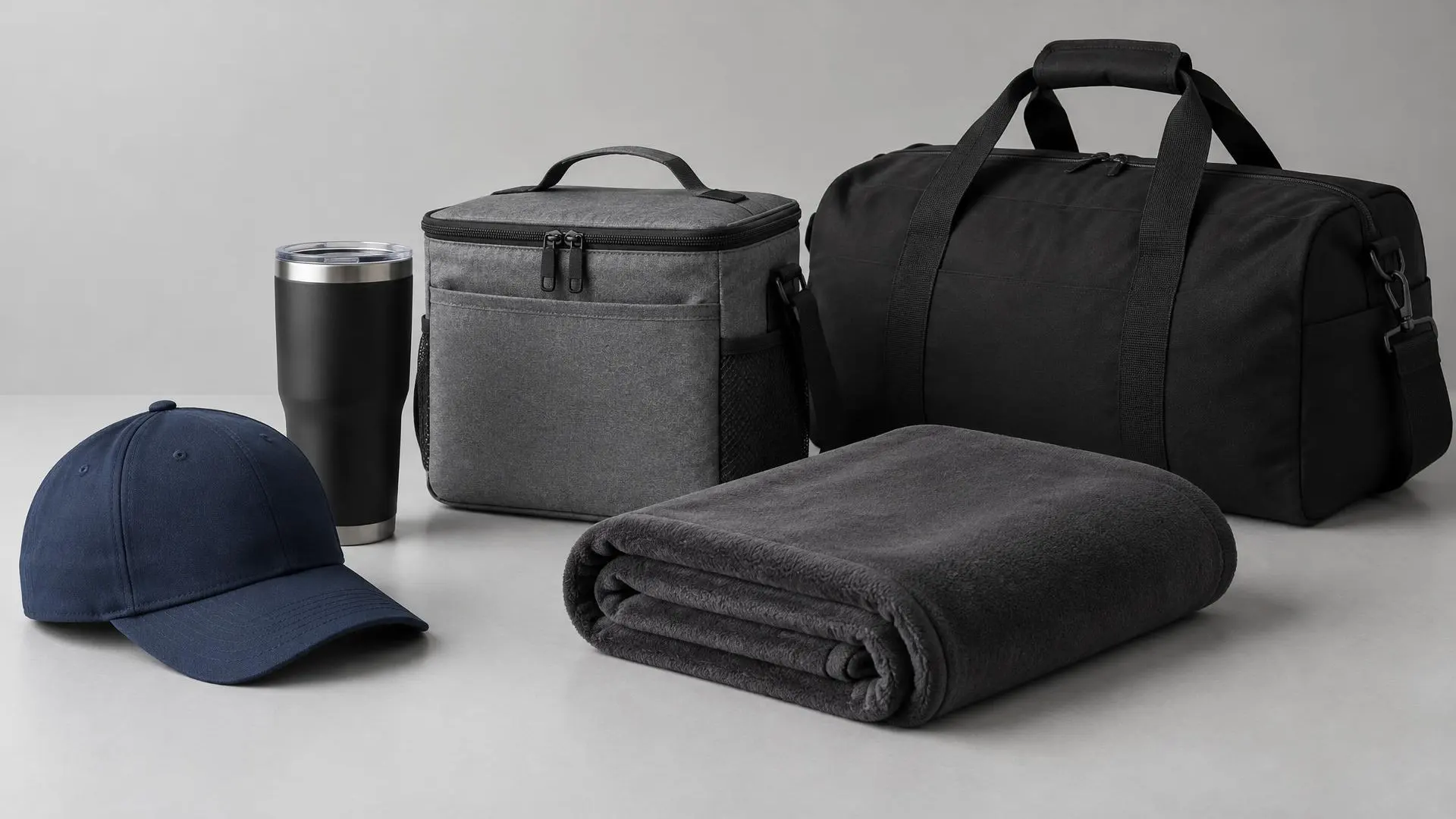 Navy cap, black tumbler, gray cooler bag, folded charcoal fleece blanket and black duffel bag on light gray.